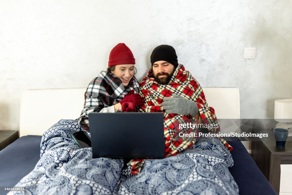 A couple wearing warm clothes is freezing at home and trying to warm up under blankets in a bedroom.