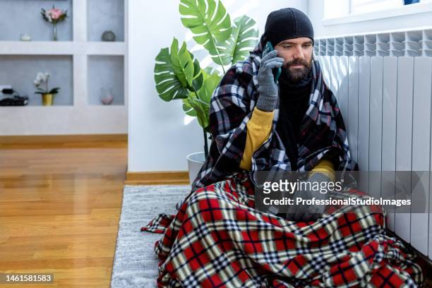 a young man is feeling displeased due to the cold temperature in his apartment sitting near a radiator while calling an energetic service. - broken air conditioner stock pictures, royalty-free photos & images