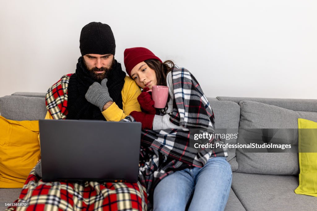 A man and woman in warm clothing are shivering and wearing a blanket and freezing at home while warming up with hot tea.