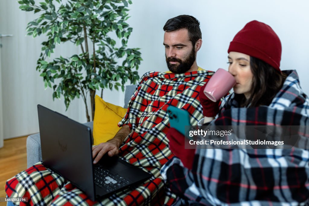 A young couple is feeling displeased due to the cold temperature in their apartment sitting on a sofa and drinking tea.