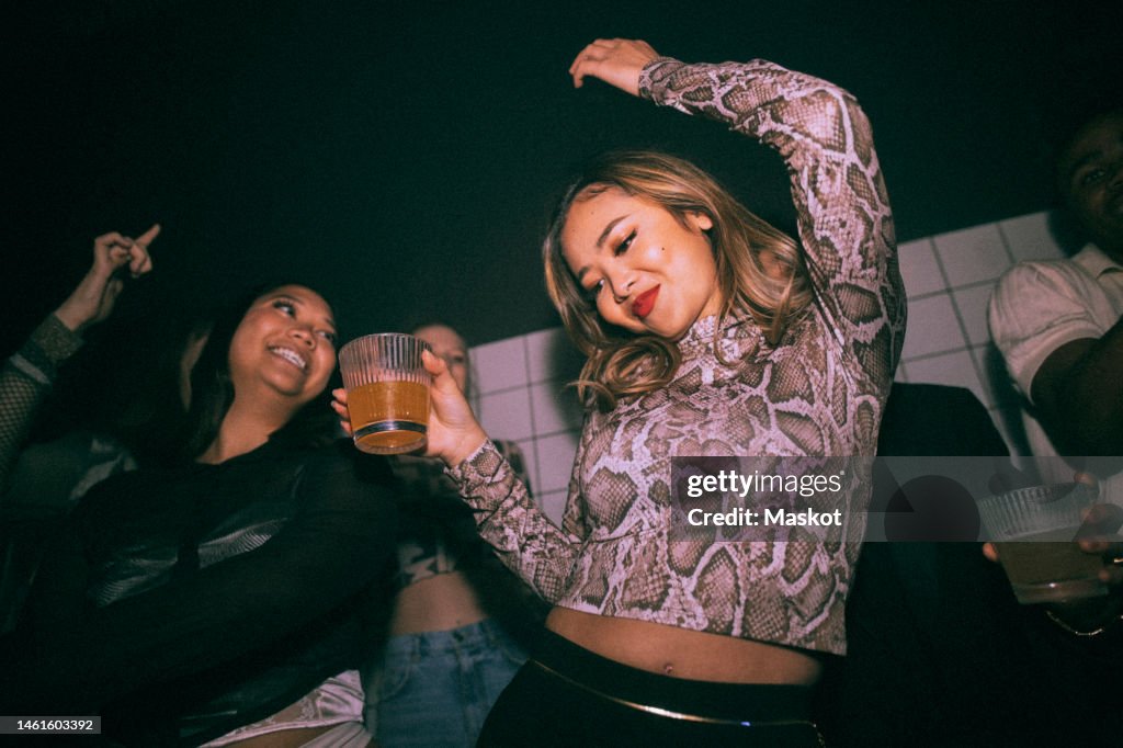 Carefree young woman with drink dancing by female friend enjoying at nightclub