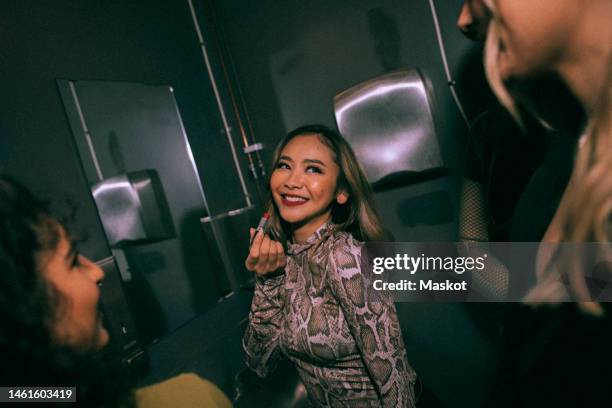 smiling young woman applying lipstick while looking at friends in nightclub bathroom - nightclub bathroom stock pictures, royalty-free photos & images