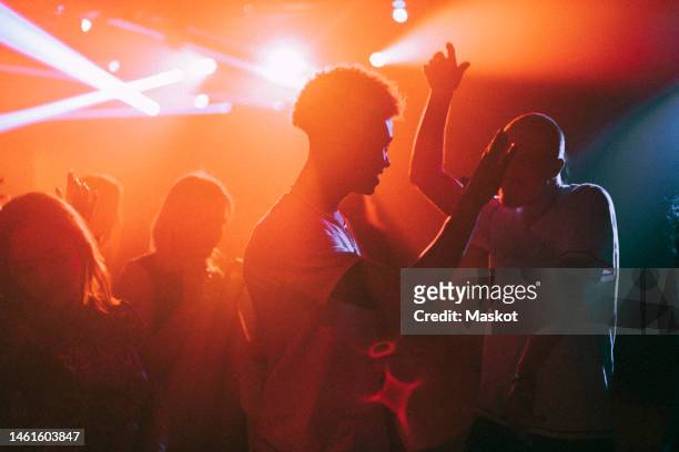young men and women dancing against illuminated red spotlights at nightclub - rave stock-fotos und bilder