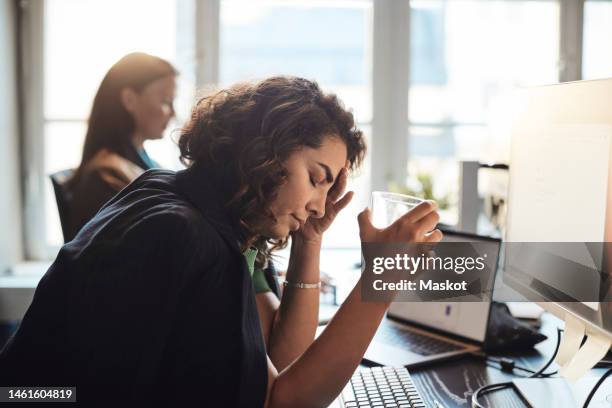 tired businesswoman with head in hand holding drinking glass at office - overwerkt stockfoto's en -beelden