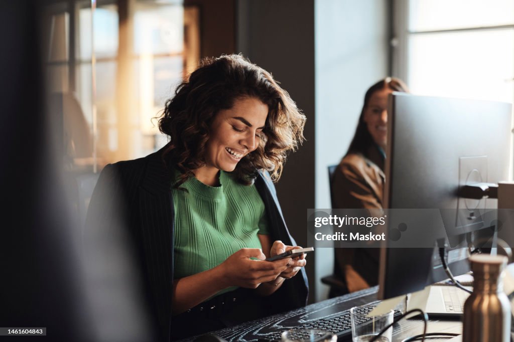 Happy female entrepreneur using mobile phone sitting at desk in office