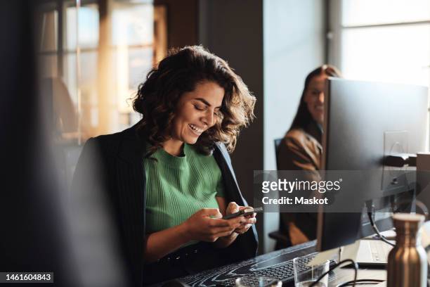 happy female entrepreneur using mobile phone sitting at desk in office - business scandinavia stock pictures, royalty-free photos & images