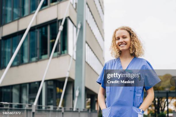 portrait of happy female nurse with hands in pockets standing in front of hospital - female nurse stock-fotos und bilder