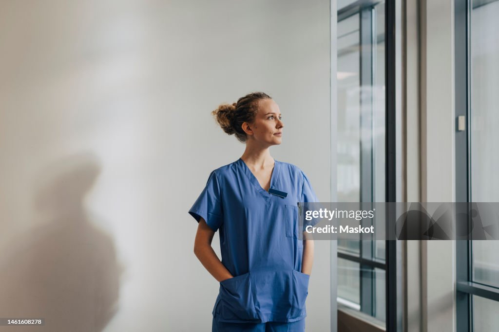 Thoughtful young nurse standing with hands in pockets against wall at hospital