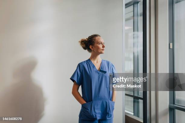 thoughtful young nurse standing with hands in pockets against wall at hospital - medical scrubs bildbanksfoton och bilder