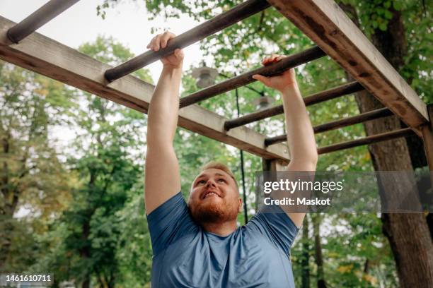 smiling overweight man practicing monkey bars at park - klimrek stockfoto's en -beelden