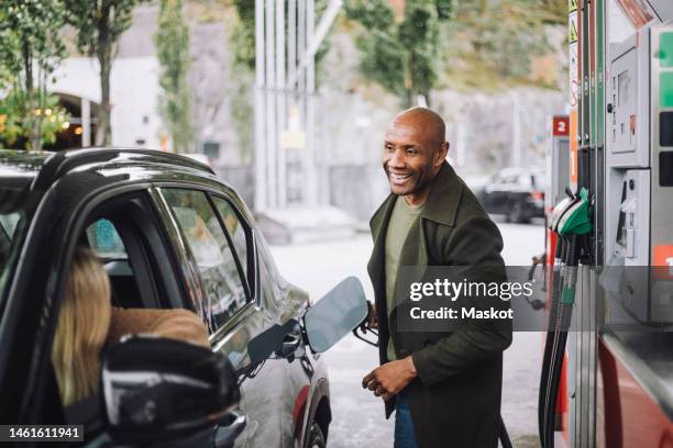 man with shaved head talking to woman while refueling car while at gas station - ガソリンスタンド ストックフォトと画像