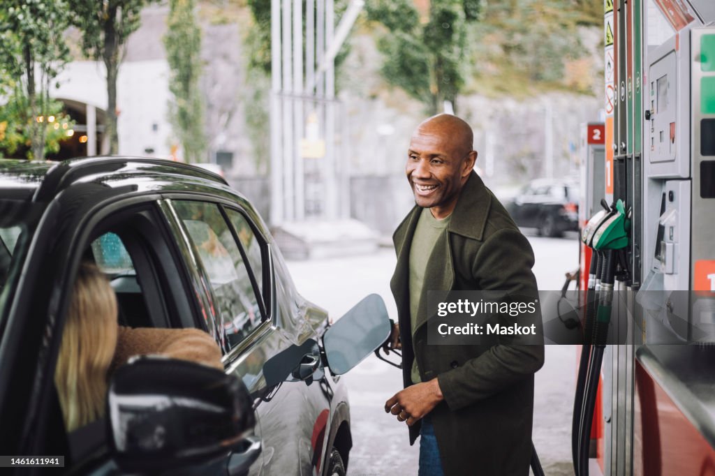 Man with shaved head talking to woman while refueling car while at gas station