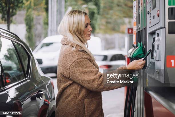 side view of blond woman doing payment via smart phone while standing at fuel station - bensinstation bildbanksfoton och bilder
