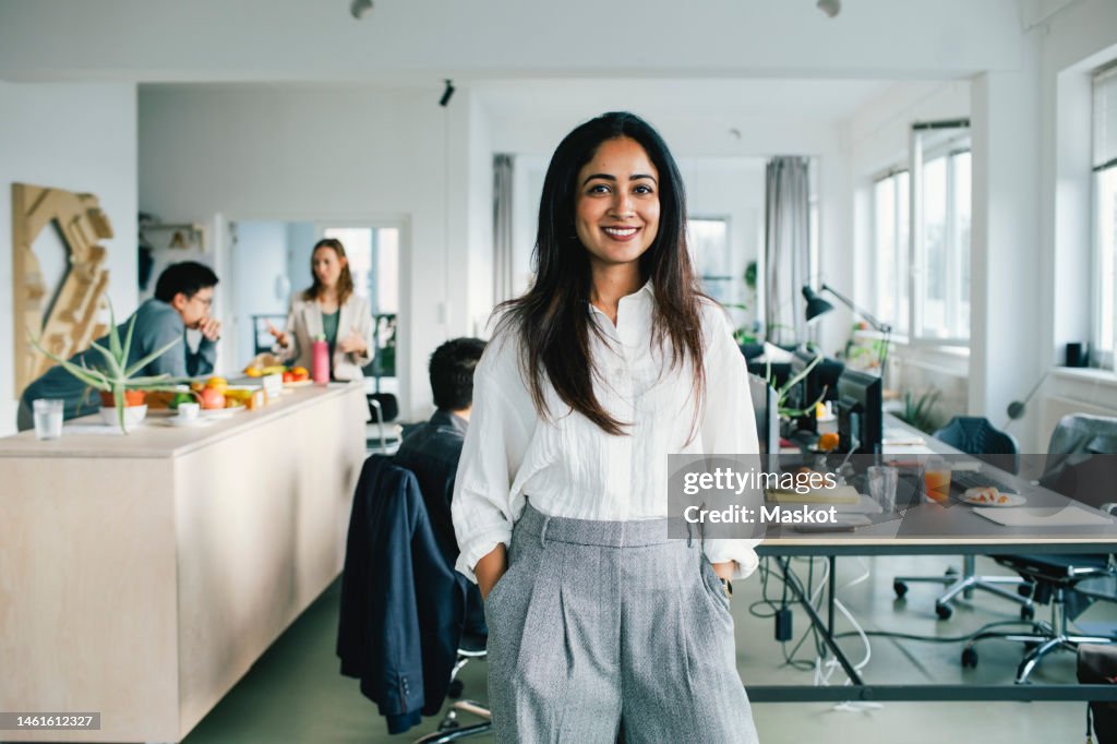 Portrait of confident smiling businesswoman standing with hands in pockets at office