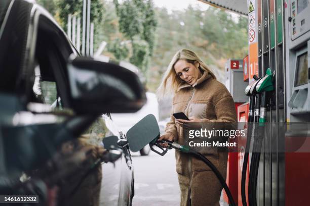 woman using mobile phone while refueling car at gas station - abastecer imagens e fotografias de stock