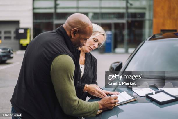 saleswoman assisting male customer while doing paperwork standing near car - car dealership stock pictures, royalty-free photos & images