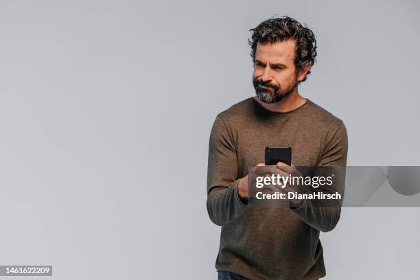 scatto in studio di un uomo maturo serio e scettico con abiti casual con il suo cellulare in mano su sfondo grigio - sospetto foto e immagini stock