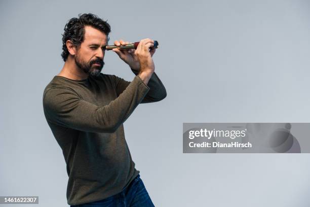 studio shot of a casual dressed mature man with natural black and gray hair looking into the distance with binoculars on grey background in direction of the copy space - knowledge is power stock pictures, royalty-free photos & images