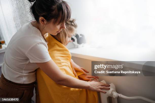 mother and daughter warming themselves from the heating radiator at home - electric fire stock pictures, royalty-free photos & images