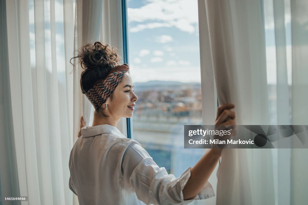 One young woman opens the curtains in the room in the morning at sunrise, she is dressed in a white shirt, ready for work and daily duties, morning routine