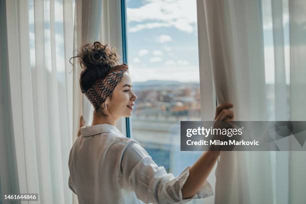 one young woman opens the curtains in the room in the morning at sunrise, she is dressed in a white shirt, ready for work and daily duties, morning routine - olhar através da janela imagens e fotografias de stock
