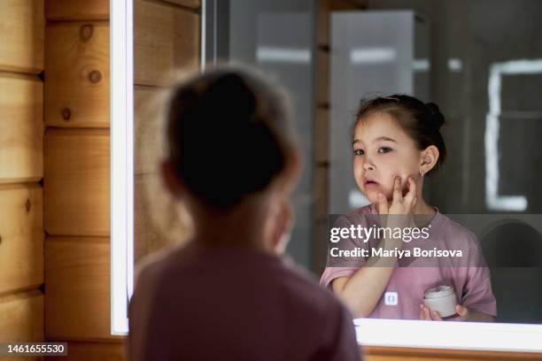 a girl in pink is standing in the bathroom in front of a illuminated mirror and smears her face with cream. - girl in mirror stockfoto's en -beelden