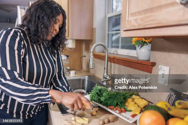 mentre una donna afroamericana nera matura e attraente prepara i pasti nella sua cucina, taglia lo zenzero e altri frutti e verdure. - "alex potemkin" or "krakozawr" foto e immagini stock