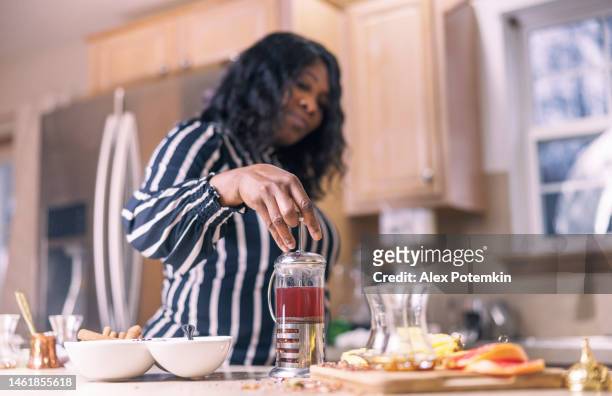 una bella donna afroamericana nera sta preparando una tisana nella sua cucina con una pressa francese. - "alex potemkin" or "krakozawr" foto e immagini stock