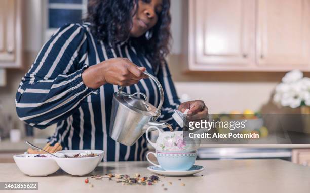 una donna afroamericana nera matura e attraente sta preparando una tisana, versando il tè da un bollitore a una teiera nella sua cucina. - "alex potemkin" or "krakozawr" foto e immagini stock