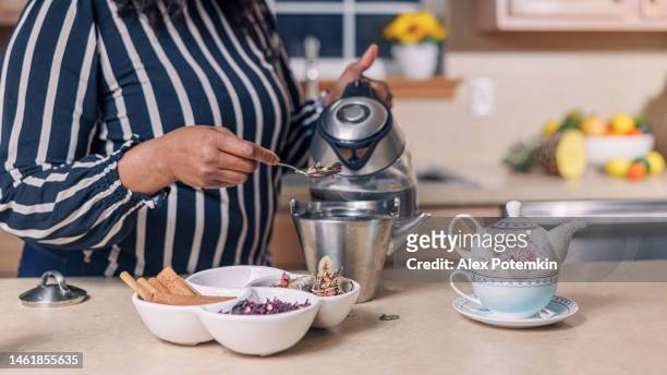 una matura e attraente donna afroamericana nera sta preparando una tisana usando un bollitore e una teiera nella sua cucina. - "alex potemkin" or "krakozawr" foto e immagini stock