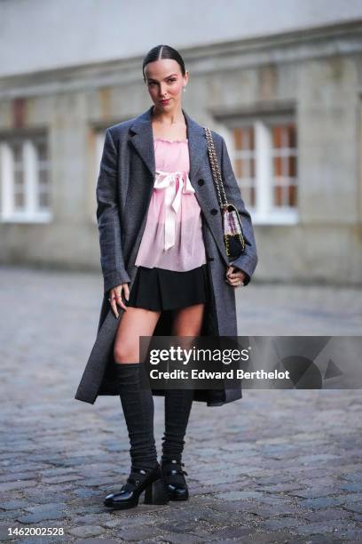 Clemence Bertrand wears silver earrings, a necklace, a pale pink ruffled silk blouse, a gray long wool coat, a black pleated short skirt, a black /...