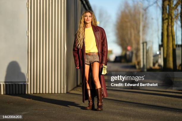 Emili Sindlev wears gold earrings, a yellow wool pullover, a burgundy shiny leather oversized long coat, beige with brown and red houndstooth print...