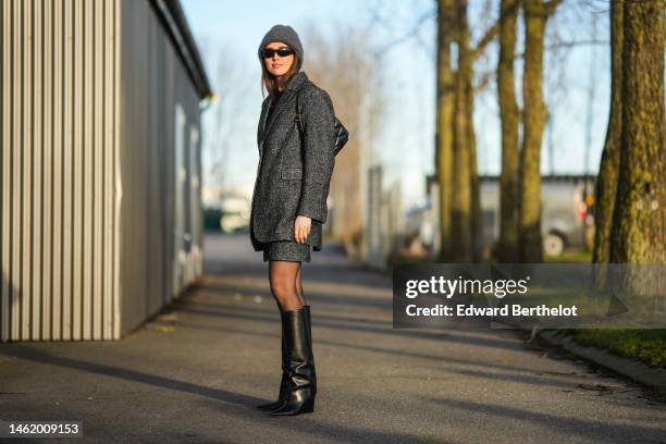 Darja Barannik wears a gray wool beanie, black sunglasses, gold earrings, a black t-shirt, a dark gray oversized blazer jacket, a matching dark gray...