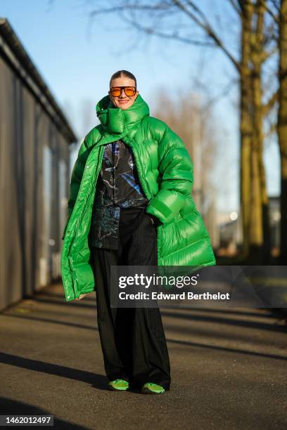 Marianne Theodorsen wears orange sunglasses, a green hoodie long oversized puffer jacket, a black with green / orange and blue print pattern shirt,...
