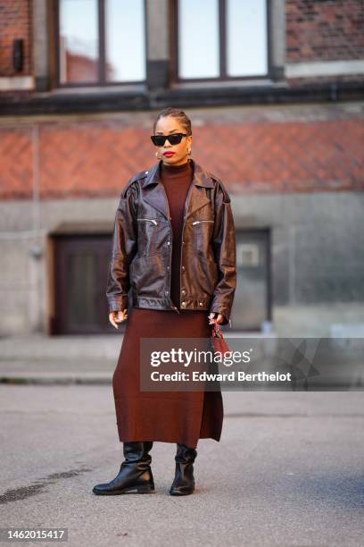 Ellie Delphine wears black sunglasses, gold earrings, a dark brown high neck long dress, a dark brown shiny varnished leather oversized biker jacket,...