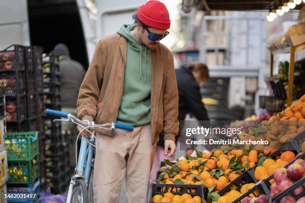trendy young guy with bike is walking near farmers market stalls choosing fresh tangerines - clementine street stock pictures, royalty-free photos & images