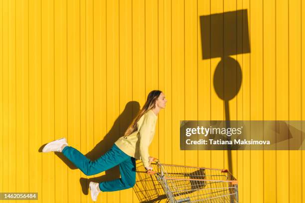 young woman with shopping cart on yellow wall background. shadow of sign stop on the wall. - shopping fun stock-fotos und bilder