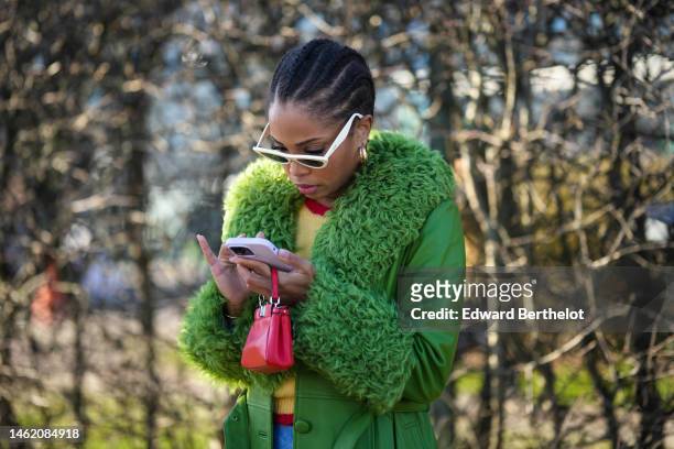 Ellie Delphine wears white latte sunglasses, gold earrings, a yellow wool with embroidered red cherry pattern pullover, a green shiny leather with...