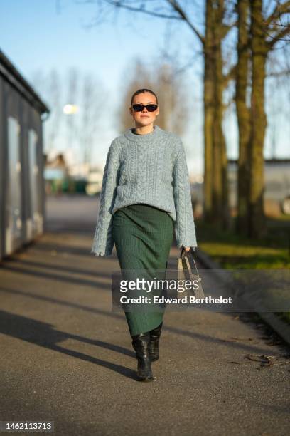 Guest wears black sunglasses, gold and white pearl pendant earrings, a pale gray braided wool pullover, a dark green ribbed wool long dress, a beige...