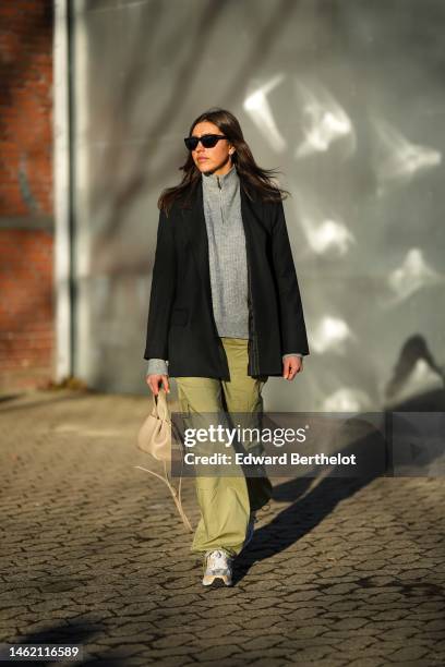 Guest wears black sunglasses, a pale gray high neck pullover, a black blazer jacket, khaki large cargo pants, a beige matte leather handbag, sneakers...