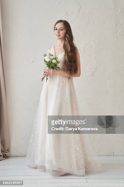 a beautiful young bride in a wedding dress and flowers standing in a room with a beautiful interior with curtains. a woman in a full-length long dress. a lifestyle with a copy space. - fabulous full lengths foto e immagini stock