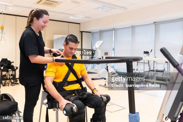 mechanical exoskeleton. physiotherapy in a modern hospital: smile of the physiotherapist woman placing the disabled person to get up with the robotic skeleton. scientists, engineers and physiotherapy rehabilitation doctors use a tablet to help - fake smile ストックフォトと画像