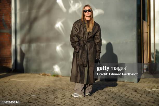 Guest wears black sunglasses, a black high neck pullover, a dark brown belted oversized coat, brown suit large pants, a black shiny leather handbag,...