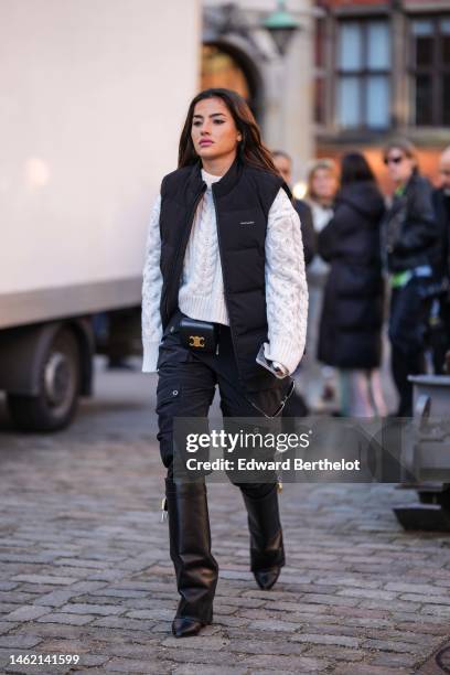 Guest wears a white braided wool pullover, a black sleeveless puffer jacket from Holzweiler, a black shiny leather belted bag from Celine,...