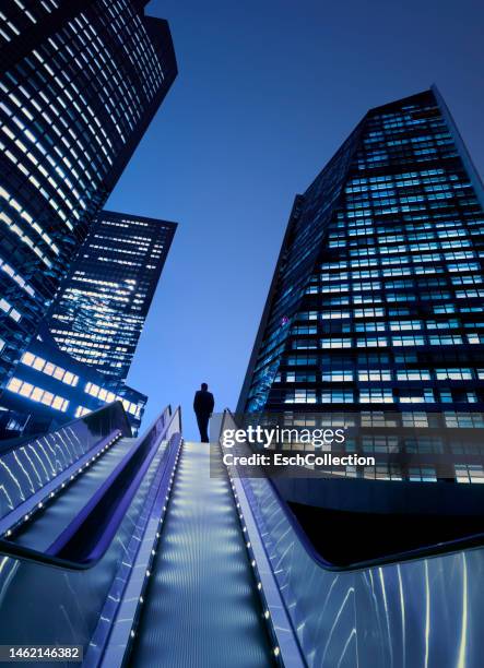 businessman on top of moving escalator at illuminated business district - escalators stock pictures, royalty-free photos & images