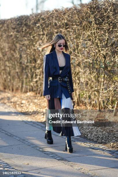 Guest wears black sunglasses, silver earrings, a silver large chain necklace, a black V-neck / cropped top, a navy blue striped print pattern blazer...