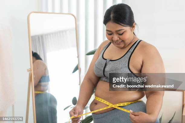 nice chubby woman measuring her waist while standing in front of the mirror - fat foto e immagini stock