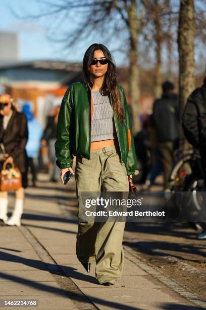 Guest wears black sunglasses, a pale gray ribbed wool t-shirt, a green shiny leather bomber coat, pale khaki large wide legs cargo pants, a brown...