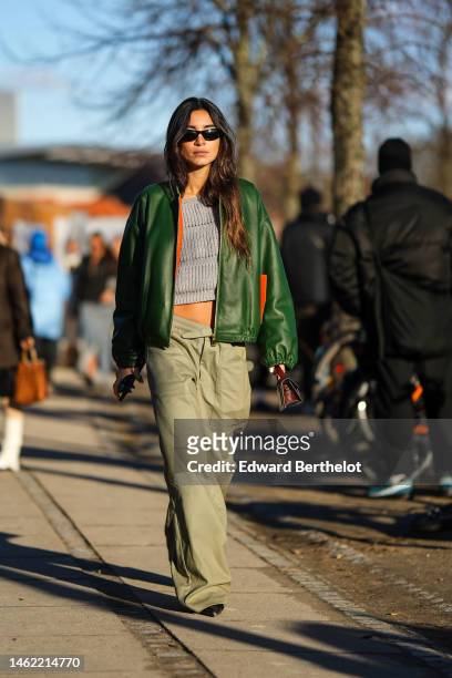 Guest wears black sunglasses, a pale gray ribbed wool t-shirt, a green shiny leather bomber coat, pale khaki large wide legs cargo pants, a brown...
