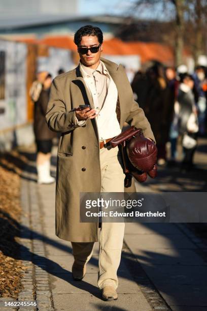 Guest wears black sunglasses, a beige shirt, a white buttoned cardigan, a beige long trench coat, a dark brown shiny leather puffy handbag, beige...